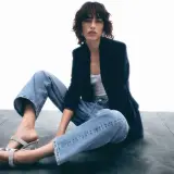 Frau sitzt lässig in Jeans und Blazer, zeigt, wie man Jeans elegant kombinieren kann. Glitzernde Schuhe runden den Look ab.
