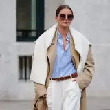 Eine Frau trägt einen beigefarbenen Trenchcoat, ein hellblaues Hemd und weiße Hose. Das Outfit zeigt, wie man ein beiges Oberteil kombinieren kann, auch für Herren.