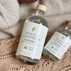 Zwei Flaschen "Wool & Cashmere" Bio-Waschmittel neben Garnknäueln und Strickwaren. Perfekt für die sanfte **Wolle waschen mit Shampoo**.