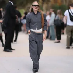 Frau mit auffälliger Sonnenbrille trägt eine graue Nadelstreifen-Jacke und eine weite anzughose zu kurz, die über ihren Schuhen endet.