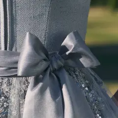 Ein Dirndl mit einer großen, grauen Schleife. Die Schleife ist Teil des Kleides, das auch mit Pailletten und Stickereien verziert ist. Perfekt für Kinder, die sich festlich kleiden möchten.