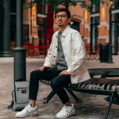 Ein Mann im smart casual Look für den Sommer sitzt auf einer Bank. Er trägt eine weiße Jacke über einem grauen T-Shirt, schwarze Jeans und weiße Sneaker.