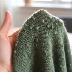 Grüner wollpullover fusselt leicht, mit kleinen Noppenmuster. Eine Hand hält den Stoff.