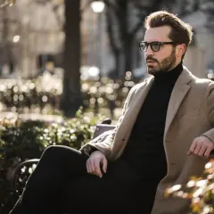 Ein Mann mit Bart und Brille sitzt stilvoll gekleidet in einem Park. Er trägt einen beigen Mantel über einem schwarzen Rollkragenpullover und dunklen Hosen.