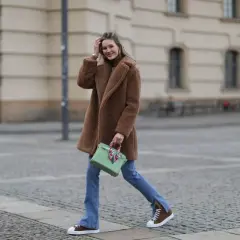 Frau in braunem Teddy-Mantel, blauer Jeans und braunen Schuhen mit weißer Sohle geht lässig über Kopfsteinpflaster.