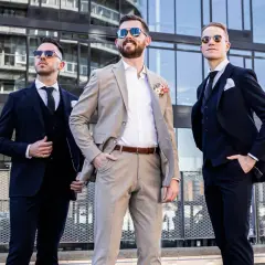Drei Männer in Anzügen vor moderner Architektur. Der mittlere Mann trägt einen hellbraunen Anzug, die anderen dunkle. Eine stilvolle Inspiration, wie man schwarze Hose kombinieren kann für eine Hochzeit.