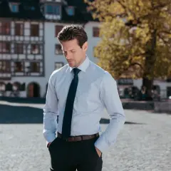Mann in einem hellen Hemd und dunkelblauem Hemd mit Krawatte, Hände in den Hosentaschen, steht auf einem Platz vor historischen Gebäuden.