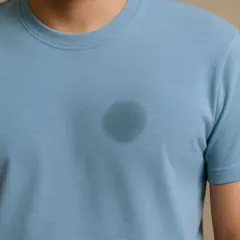 Ein dunkler Fleck auf einem hellblauen Baumwoll-T-Shirt, der zeigt, wie man Fettflecken aus Baumwolle entfernen kann.