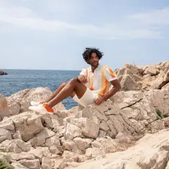 Mann sitzt auf Felsen am Meer, trägt ein buntes Poloshirt, weiße kurze Hose und passende schuhe zu kurzen hosen herren mit orangen Akzenten.