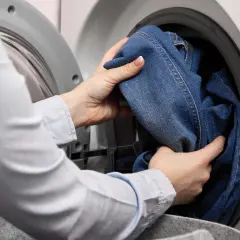 Hände legen Jeans in die Waschmaschine, um sie mit anderen Kleidungsstücken zu waschen.