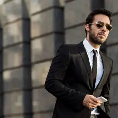 Ein Mann im Anzug mit Sonnenbrille hält ein Smartphone. Er steht vor einer modernen Gebäudefassade.
