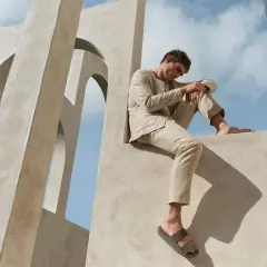Mann in einem hellen, beigen Anzug mit einem blauen Sakko und einer beigen Hose sitzt auf einer Betonstruktur vor blauem Himmel.