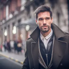 Ein Mann im eleganten Trenchcoat Herren Style, mit Bart und dunklem Haar, blickt nachdenklich in die Ferne.