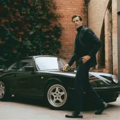 Ein Mann im eleganten Anzug mit Stiefel lehnt sich an einen klassischen Porsche.