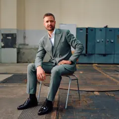 Mann in grauem Anzug mit Sakko mit 2 Schlitzen hinten, sitzt auf einem Stuhl in einer Industriehalle.