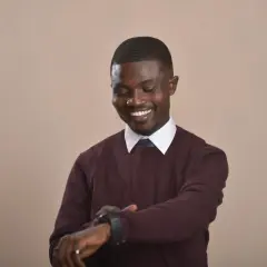 Ein Mann mit dunkler Haut und einem Pullover über Hemd und Krawatte schaut auf seine Uhr.
