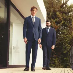 Zwei Männer in eleganten einreiher Anzügen stehen auf einer Holzterrasse vor einem modernen Gebäude.