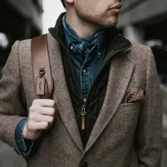 Mann im Tweed-Sakko mit Jeanshemd und Bandana, perfekt für den dresscode chic herren. Braune Ledertasche mit Coach-Anhänger.