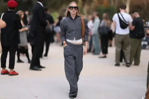Frau mit auffälliger Sonnenbrille trägt eine graue Nadelstreifen-Jacke und eine weite anzughose zu kurz, die über ihren Schuhen endet.