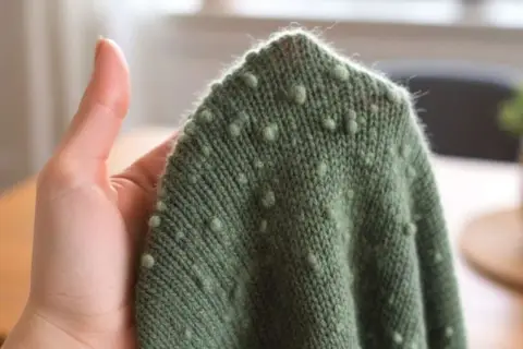 Grüner wollpullover fusselt leicht, mit kleinen Noppenmuster. Eine Hand hält den Stoff.