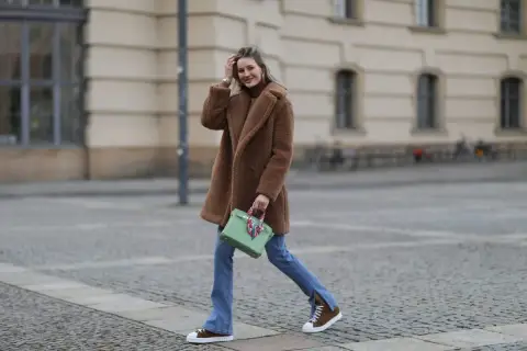 Frau in braunem Teddy-Mantel, blauer Jeans und braunen Schuhen mit weißer Sohle geht lässig über Kopfsteinpflaster.