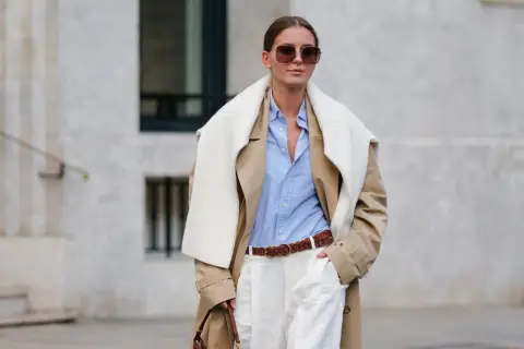 Eine Frau trägt einen beigefarbenen Trenchcoat, ein hellblaues Hemd und weiße Hose. Das Outfit zeigt, wie man ein beiges Oberteil kombinieren kann, auch für Herren.