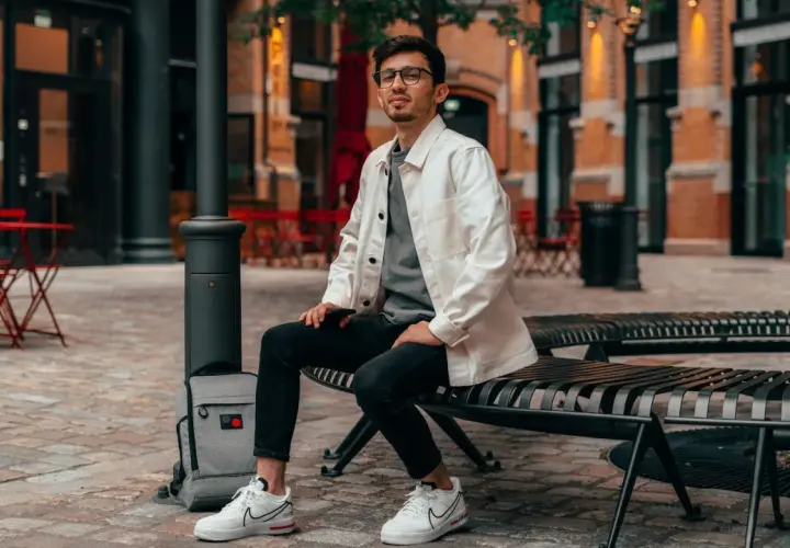 Ein Mann im smart casual Look für den Sommer sitzt auf einer Bank. Er trägt eine weiße Jacke über einem grauen T-Shirt, schwarze Jeans und weiße Sneaker.