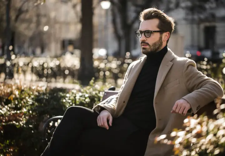 Ein Mann mit Bart und Brille sitzt stilvoll gekleidet in einem Park. Er trägt einen beigen Mantel über einem schwarzen Rollkragenpullover und dunklen Hosen.