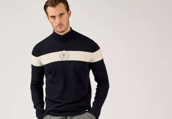 Mann in dunkelblauem Rollkragenpullover mit breitem cremefarbenem Streifen und Emblem. So kann man Rollkragenpullover kombinieren, Herren-Style.
