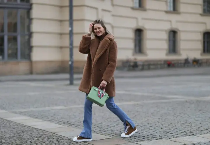 Frau in braunem Teddy-Mantel, blauer Jeans und braunen Schuhen mit weißer Sohle geht lässig über Kopfsteinpflaster.