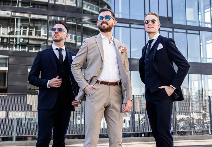 Drei Männer in Anzügen vor moderner Architektur. Der mittlere Mann trägt einen hellbraunen Anzug, die anderen dunkle. Eine stilvolle Inspiration, wie man schwarze Hose kombinieren kann für eine Hochzeit.