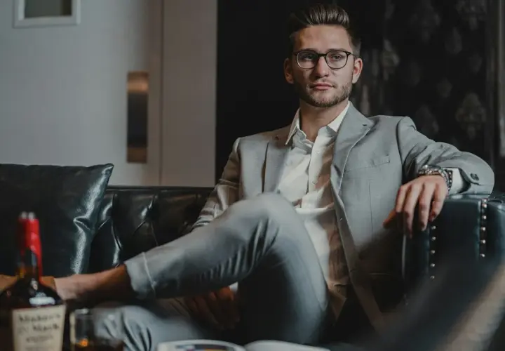 Junger Mann in stilvoller Business-Kleidung für Herren, entspannt auf einem Ledersofa mit einem Glas Whiskey.
