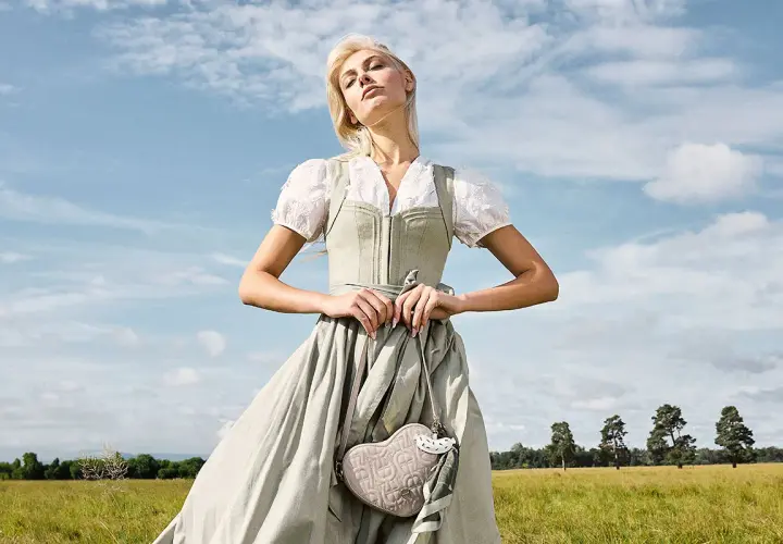 Eine Frau im Dirndl mit zarten Dirndl Farben steht auf einer grünen Wiese. Sie hält eine herzförmige Tasche.