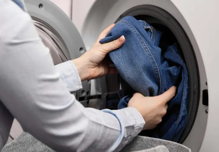 Hände legen Jeans in die Waschmaschine, um sie mit anderen Kleidungsstücken zu waschen.