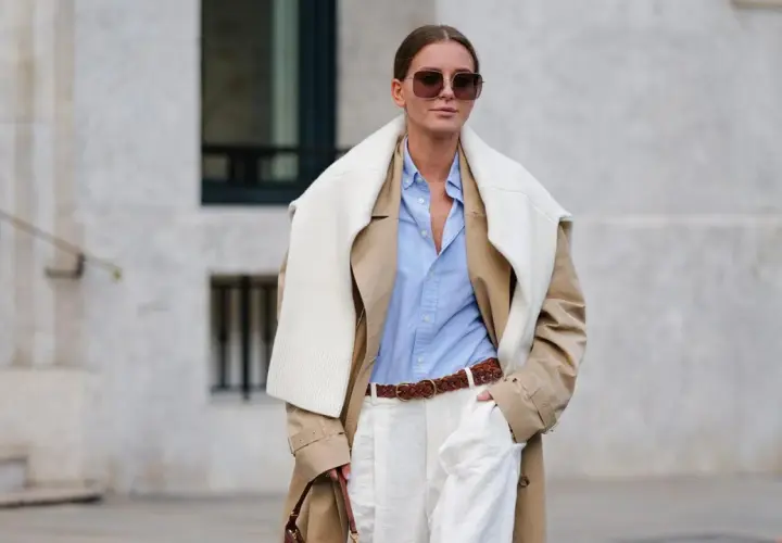 Eine Frau trägt einen beigefarbenen Trenchcoat, ein hellblaues Hemd und weiße Hose. Das Outfit zeigt, wie man ein beiges Oberteil kombinieren kann, auch für Herren.