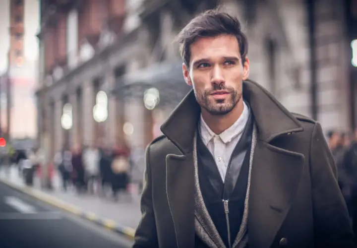 Ein Mann im eleganten Trenchcoat Herren Style, mit Bart und dunklem Haar, blickt nachdenklich in die Ferne.