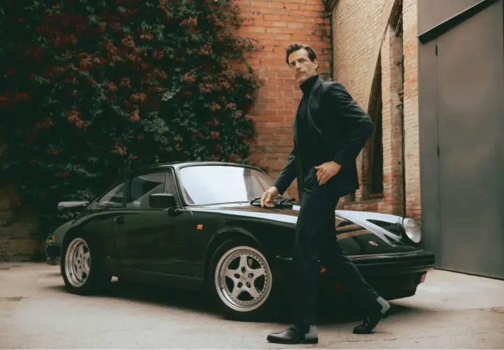 Ein Mann im eleganten Anzug mit Stiefel lehnt sich an einen klassischen Porsche.