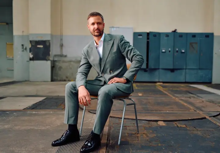 Mann in grauem Anzug mit Sakko mit 2 Schlitzen hinten, sitzt auf einem Stuhl in einer Industriehalle.