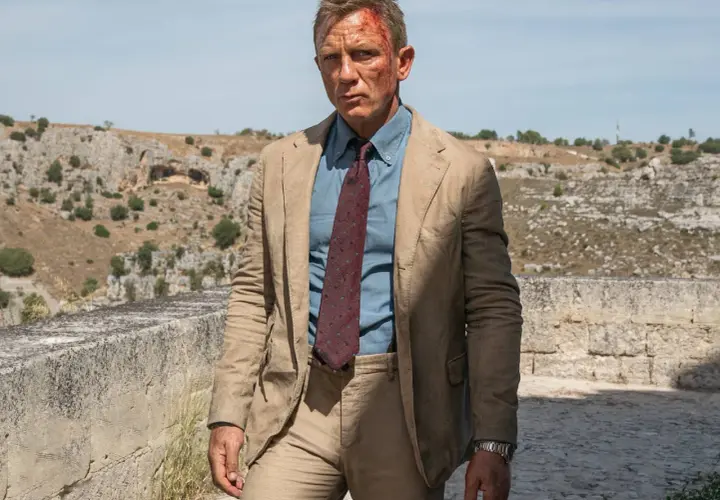 Daniel Craig in einem hellbraunen Anzug, einem blauen Hemd und einer gemusterten Krawatte, typisch für James Bond Outfits, mit leichten Verletzungen im Gesicht.