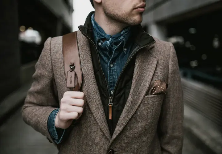 Mann im Tweed-Sakko mit Jeanshemd und Bandana, perfekt für den dresscode chic herren. Braune Ledertasche mit Coach-Anhänger.