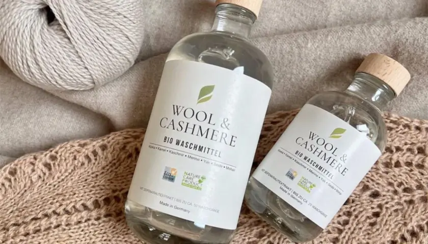 Zwei Flaschen "Wool & Cashmere" Bio-Waschmittel neben Garnknäueln und Strickwaren. Perfekt für die sanfte **Wolle waschen mit Shampoo**.