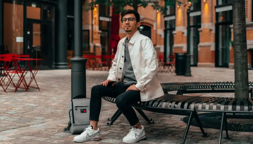 Ein Mann im smart casual Look für den Sommer sitzt auf einer Bank. Er trägt eine weiße Jacke über einem grauen T-Shirt, schwarze Jeans und weiße Sneaker.