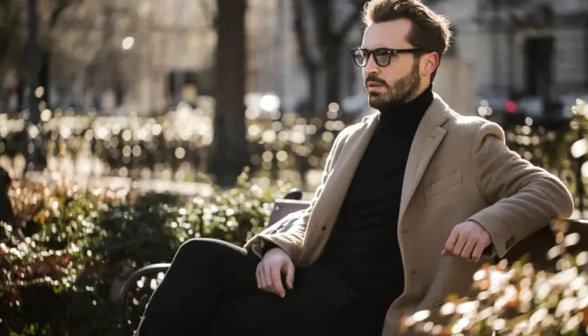 Ein Mann mit Bart und Brille sitzt stilvoll gekleidet in einem Park. Er trägt einen beigen Mantel über einem schwarzen Rollkragenpullover und dunklen Hosen.