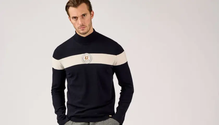 Mann in dunkelblauem Rollkragenpullover mit breitem cremefarbenem Streifen und Emblem. So kann man Rollkragenpullover kombinieren, Herren-Style.