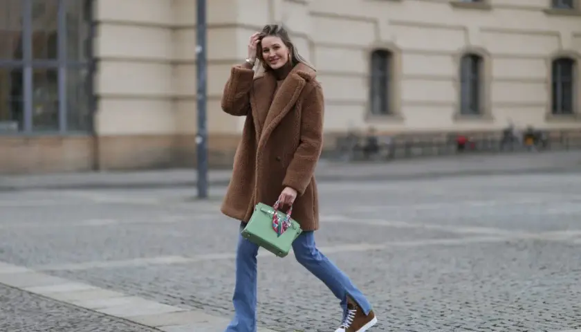 Frau in braunem Teddy-Mantel, blauer Jeans und braunen Schuhen mit weißer Sohle geht lässig über Kopfsteinpflaster.