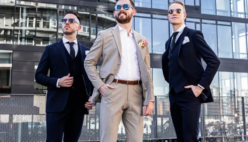 Drei Männer in Anzügen vor moderner Architektur. Der mittlere Mann trägt einen hellbraunen Anzug, die anderen dunkle. Eine stilvolle Inspiration, wie man schwarze Hose kombinieren kann für eine Hochzeit.