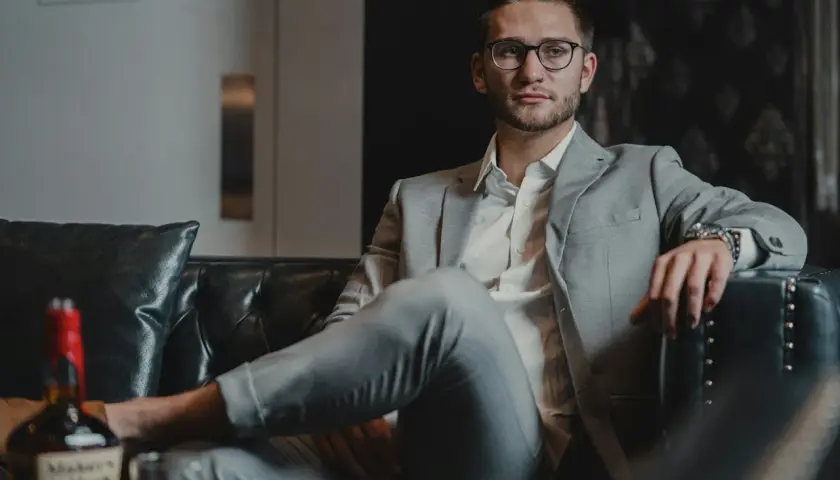 Junger Mann in stilvoller Business-Kleidung für Herren, entspannt auf einem Ledersofa mit einem Glas Whiskey.