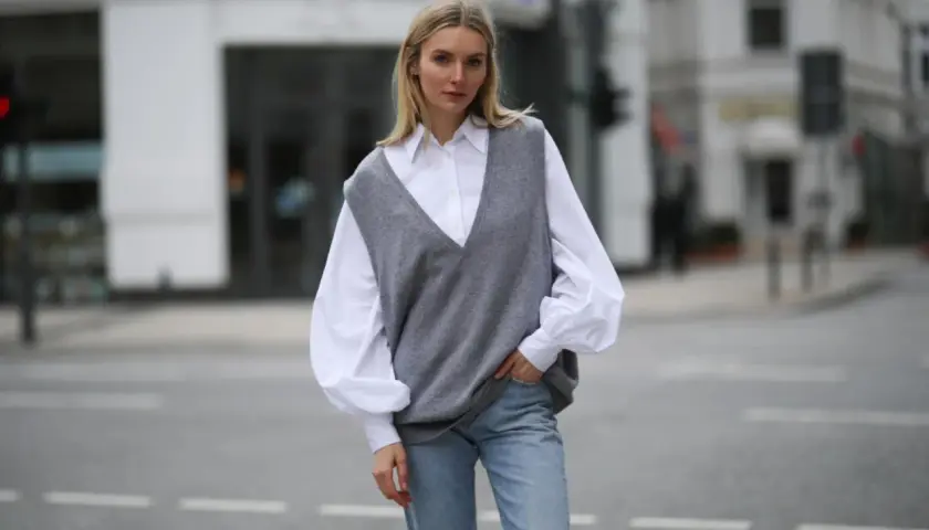 Eine Frau in einem weißen Hemd und Jeans trägt einen grauen Strick-Pullunder. So kann man Pullunder Damen kombinieren für einen lässigen Look.