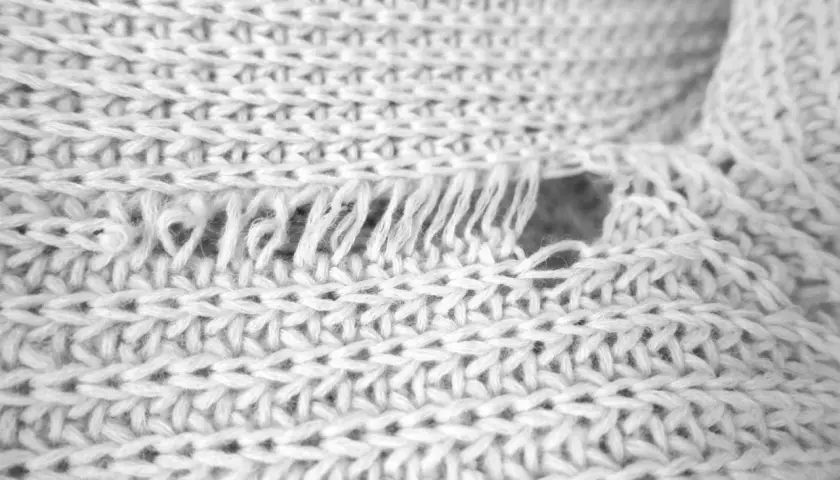 Nahaufnahme eines verfilzten und eingelaufenen Pullovers. Die Strickstruktur ist sichtbar, mit ausgefransten Fäden und einem Loch.