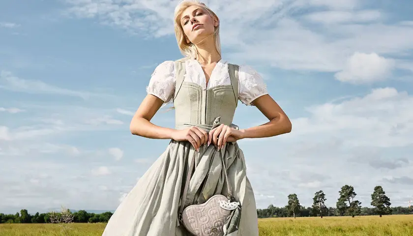 Eine Frau im Dirndl mit zarten Dirndl Farben steht auf einer grünen Wiese. Sie hält eine herzförmige Tasche.